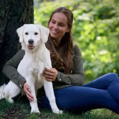 Uśmiechnięta kobieta siedzi przy drzewie, przytulając swojego białego psa.