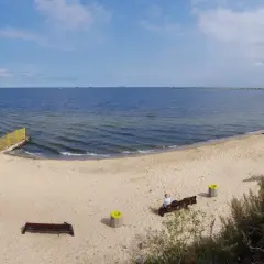 Spokojna plaża dla psów z piaskiem i morzem. W oddali widać falochron i statki.