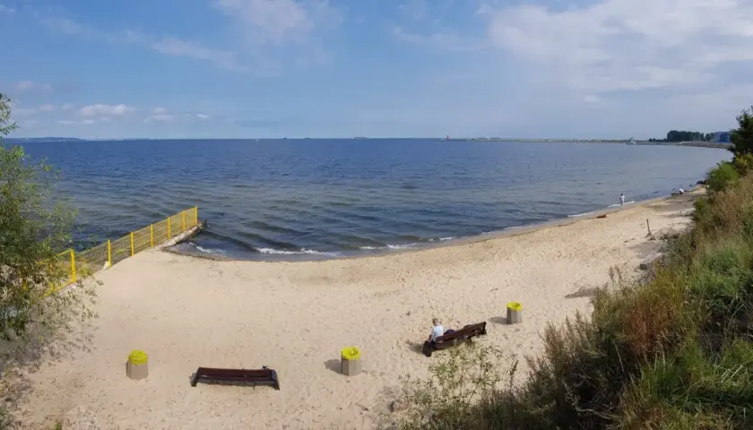 Spokojna plaża dla psów z piaskiem i morzem. W oddali widać falochron i statki.